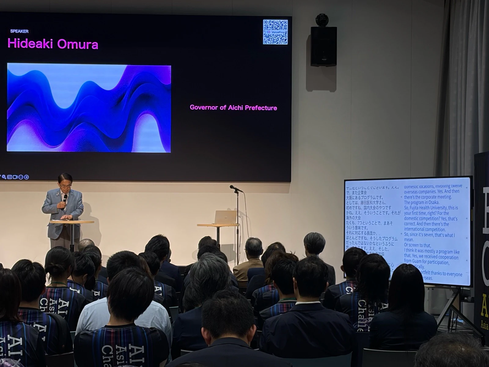 AI×MedTechで登壇する愛知県知事の大村秀章氏。メイン画面にはVoicePingのQRコード、隣の画面には日英のリアルタイム字幕が表示されている。