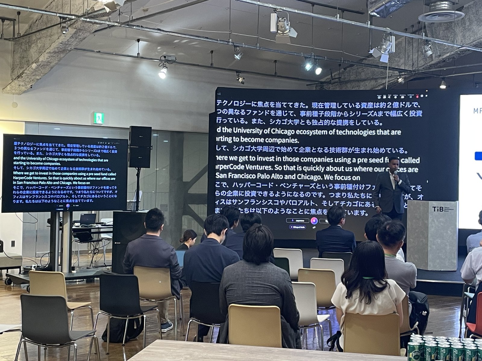 Tokyo Innovation Base でフォーラムに耳を傾ける来場者。会場のスクリーンには登壇者のスライドと VoicePing のリアルタイム翻訳の両方が映っている。