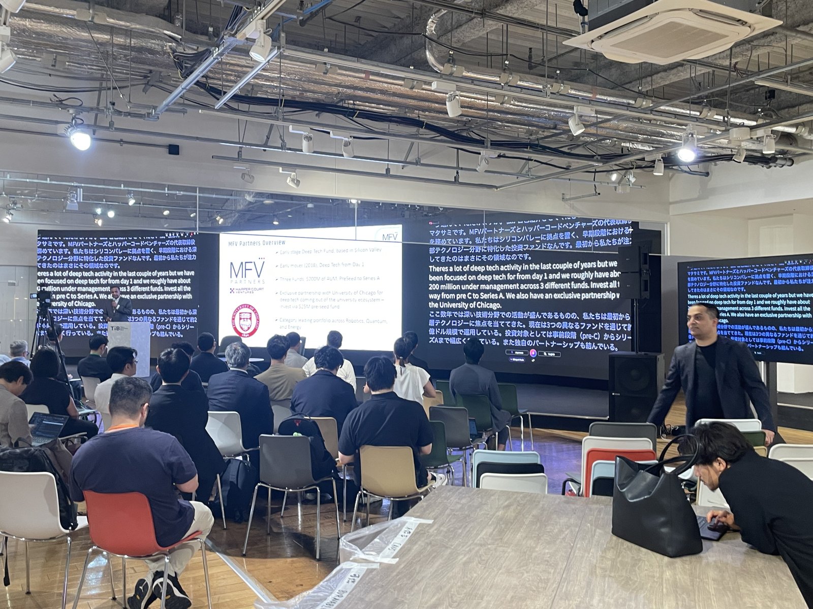 Tokyo Innovation Base の会場を広く写した一枚。来場者が2つの大型スクリーンの方を向いて着席し、片方に登壇者のスライド、もう片方に VoicePing の日英リアルタイム翻訳が表示されている。