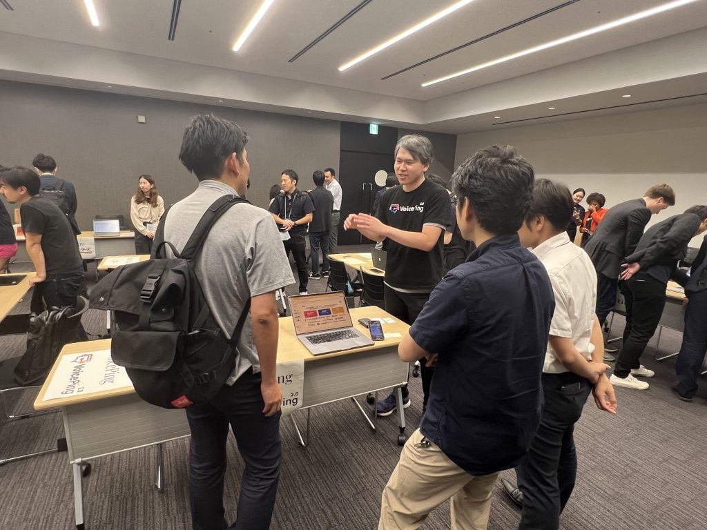 VoicePingのスタッフが、ノートPCとスマートフォンを置いた机の前で来場者に説明する様子