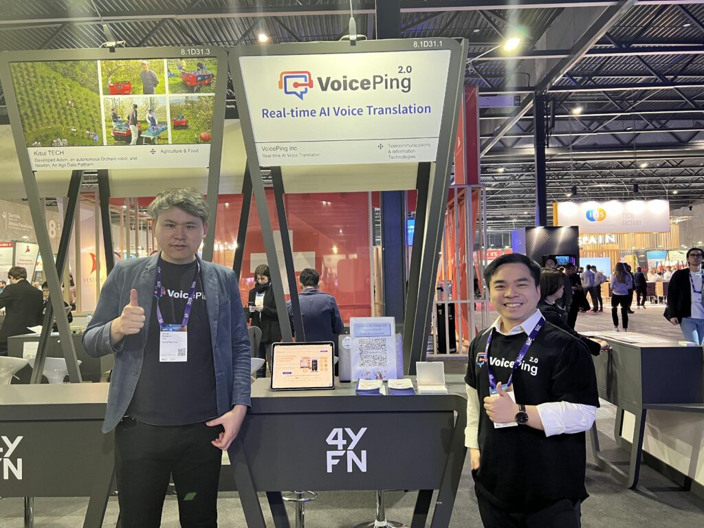 4YFNのVoicePing展示ブースで、リアルタイムAI音声翻訳の看板と並んで立つメンバー2名