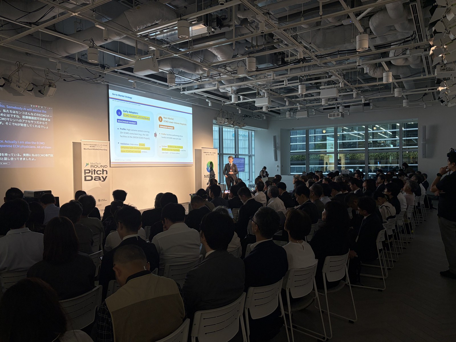 MedTech ROUND Pitch Day のメインステージで英語のピッチが行われ、その隣の2つ目のスクリーンに日本語へのリアルタイム翻訳が映し出されている様子。