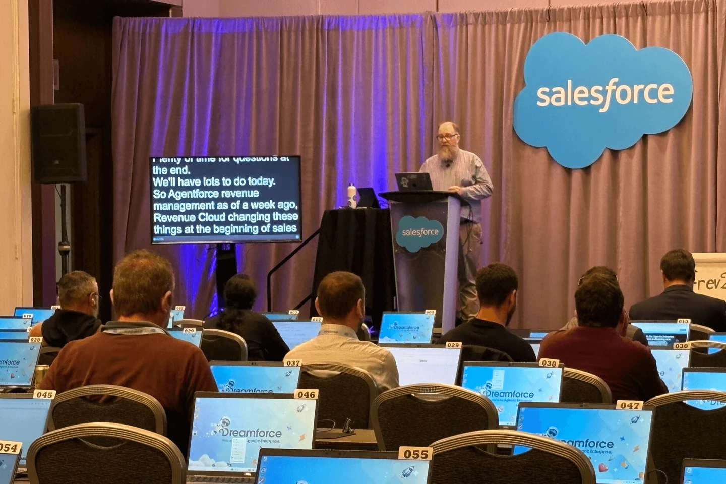 ライブ字幕モニターの横で話すカンファレンスの登壇者。着席した参加者とSalesforceのブランド表示が見える。
